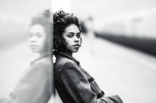 Black and white portrait of charming African American female in the coat leaning against a mirror wall; the greyscale shot of a cute black teenage girl sitting on the bench of an underground station