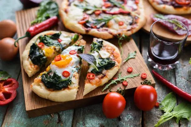 Homemade pizza and wine party. Pizzas with eggs, spinach and chili on oak chopping board over rustic blue background closeup