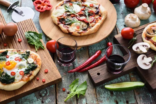Homemade pizza party. Pizzas with eggs, spinach, mushrooms and chili served with raw vegetables and red wine on dark oak chopping boards over blue rustic background closeup