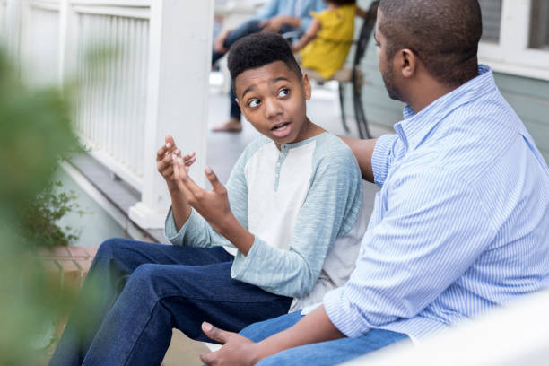 Confused preteen son talks to father on front porch A confused preteen boy sits on his front porch steps with his father and gestures in frustration.  His unrecognizable father puts an arm around his shoulders as they talk. information overload photos stock pictures, royalty-free photos & images