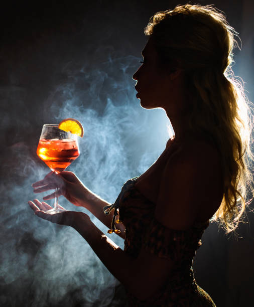 istock Beautiful young woman profile in backlight drinking Spritz isolated black background with smoke. Club party menu concept 944360582