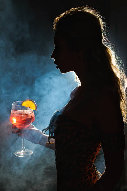 istock Beautiful young woman profile in backlight drinking Spritz isolated black background with smoke. Club party menu concept 944360746