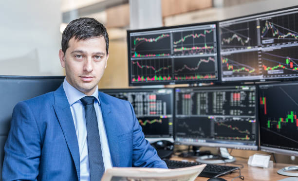 Business portrait of stock broker in traiding office. Business portrait of confident stocks broker holding a business newspaper in traiding office with multiple computer screens full of index charts and data analyses. stock certificate growth price market stock pictures, royalty-free photos & images