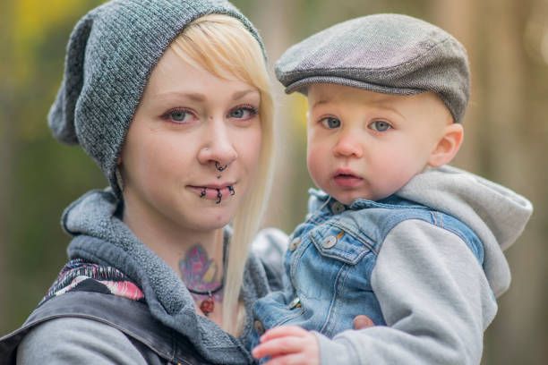 A single young mother with piercings and tattoos is standing with her baby son in her arms. They both look at the camera, she smiles. They stand in a park in the fall.