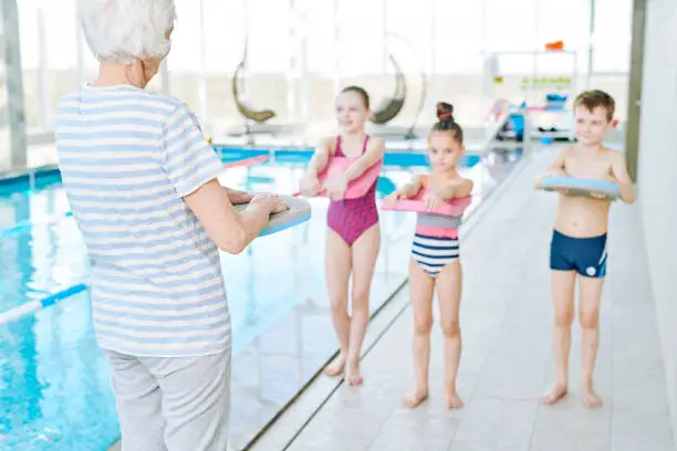 Rear view of gray-haired swimming trainer showing how swim with kickboard on land and explaining technique to kids