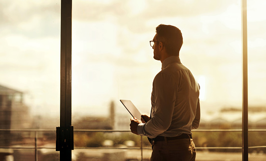 Silhouette of a mature businessman standing in front of an office window and using a digital tablet
