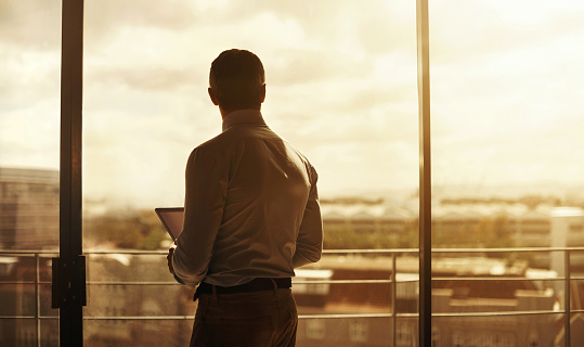 Silhouette of a mature businessman standing in front of an office window and using a digital tablet