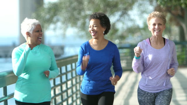 A group of three multi-ethnic mature and senior women exercising outdoors together on a city waterfront on a sunny day. They are smiling and talking as they power walk, moving toward the camera.