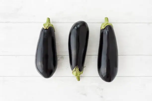 Top view of three whole aubergines on white wood background.
