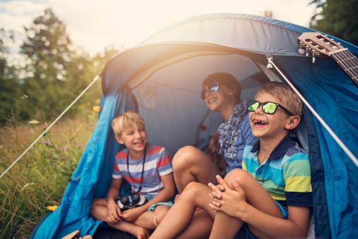 Little girl and her brothers are camping in a blue tent on the forest meadow. Kids aged 7 and 11 are laughing happily sitting inside of blue tent. Sunny summer day.\nNikon D810