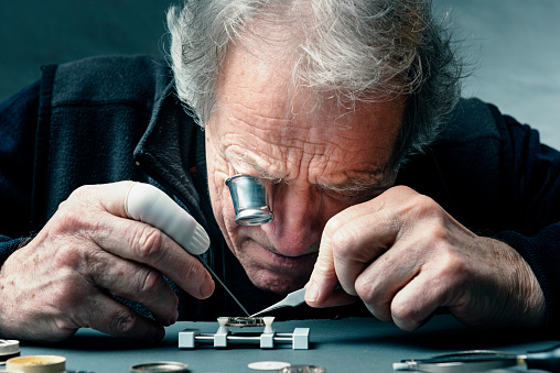 Close up portrait of a watchmaker at work as he carefully repairs the mechanism of a watch. The watch movement is held is a special vice as he works, he is using a magnifying eyepiece to see close up what he is doing. Colour, horizontal with some copy space.