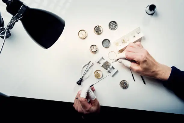 Overhead view of a watchmaker at work as he carefully repairs the mechanism of a watch. The watch movement is held is a special vice as he works. H wearing latex “finger gloves” to stop dust and dirt getting into the delicate watch mechanism. Colour, horizontal with some copy space.