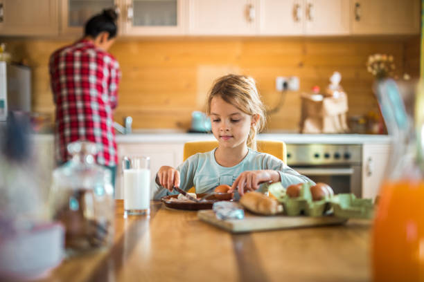 Small girl sitting at dining table and eating her breakfast. Her mother is in the background.