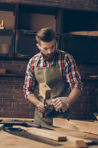 istock concenrated handsome cabinetmaker work with chisel and hammer. Stylish craftsman with brutal hairstyle and glasses at his workstation 944600212