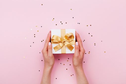 Womans hands holding gift or present box decorated confetti on pink pastel table top view. Flat lay composition for birthday or wedding.