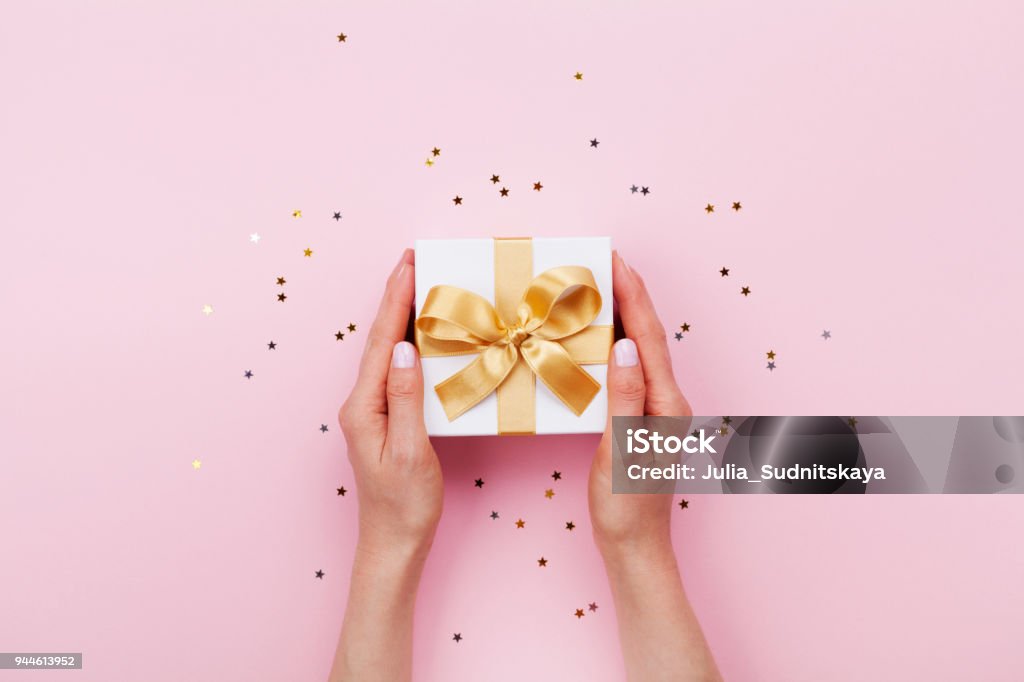 Womans hands holding gift or present box decorated confetti on pink pastel table top view. Birthday or wedding template or mockup. Womans hands holding gift or present box decorated confetti on pink pastel table top view. Flat lay composition for birthday or wedding. Gift Stock Photo