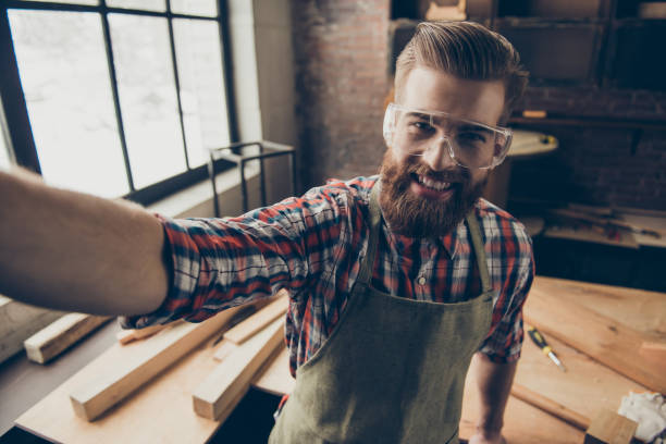 Happy successful handsome cabinetmaker make selfie photo. Stylish young entrepreneur with brutal hairstyle and glasses smiling and photographing at his workstation. Love job Happy successful handsome cabinetmaker make selfie photo. Stylish young entrepreneur with brutal hairstyle and glasses smiling and photographing at his workstation. Love job carpenter carpentry craftsperson carving stock pictures, royalty-free photos & images