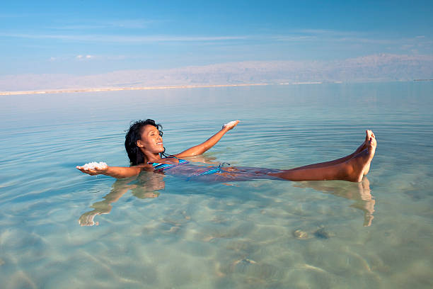 Floating in Sea of Salt. Young woman floating  on back in Dead Sea, holding mineral salt in both hands. The unusual buoyancy caused by high salinity. dead sea stock pictures, royalty-free photos & images