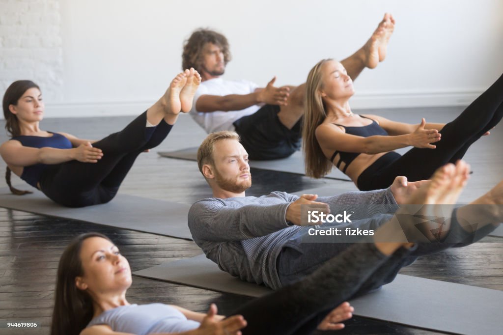 Group of young sporty people in Paripurna Navasana pose Group of young sporty people practicing yoga lesson, doing Paripurna Navasana exercise, boat pose, working out, indoor close up, studio. Healthy lifestyle concept Pilates Stock Photo