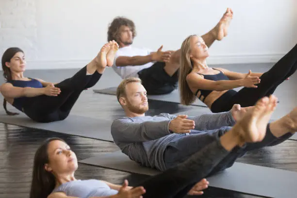 Group of young sporty people practicing yoga lesson, doing Paripurna Navasana exercise, boat pose, working out, indoor close up, studio. Healthy lifestyle concept