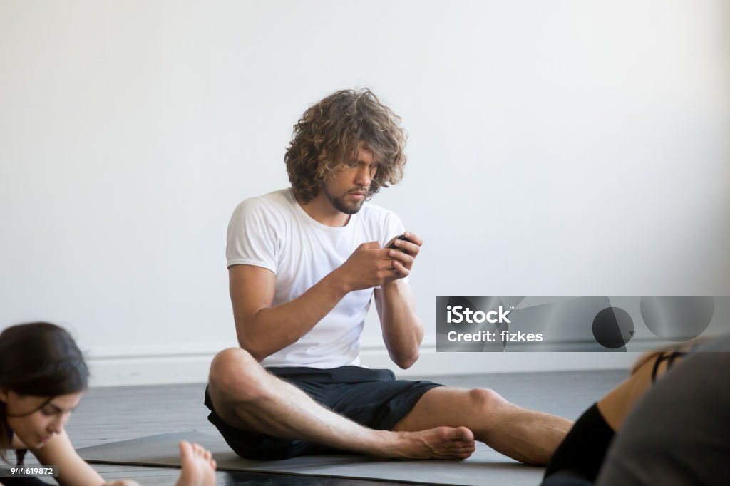 Young man sitting texting a message using his mobile phone Bored young man sitting texting a message using his mobile phone, having a break while group of young sporty people practicing yoga lesson, sitting not working out, indoor close up, yoga studio club Yoga Stock Photo