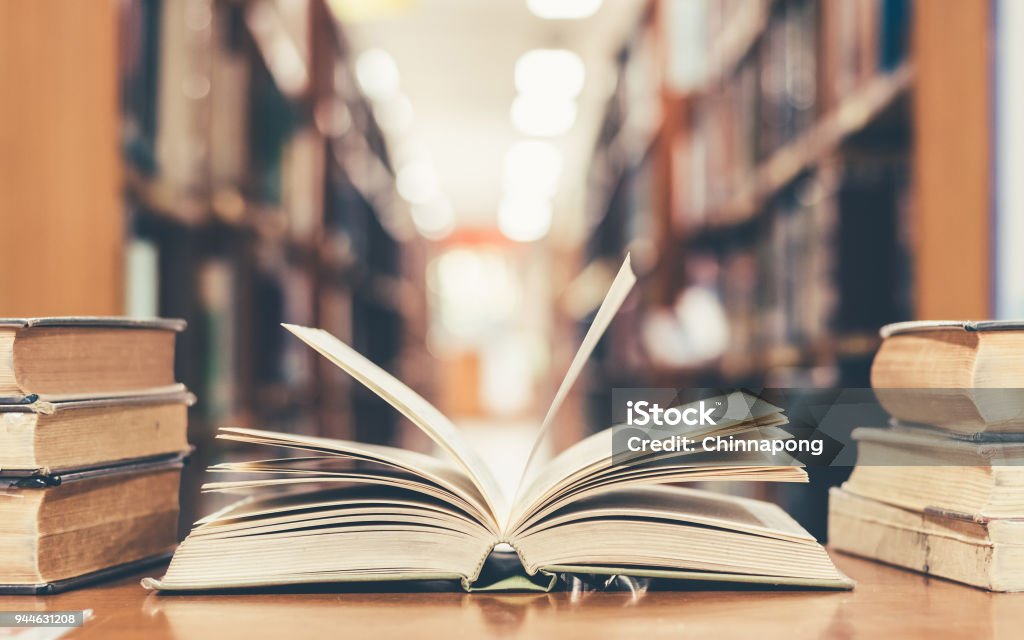 Education concept with book in library Book in library with old open textbook, stack piles of literature text archive on reading desk, and aisle of bookshelves in school study class room background for academic education learning concept Book Stock Photo
