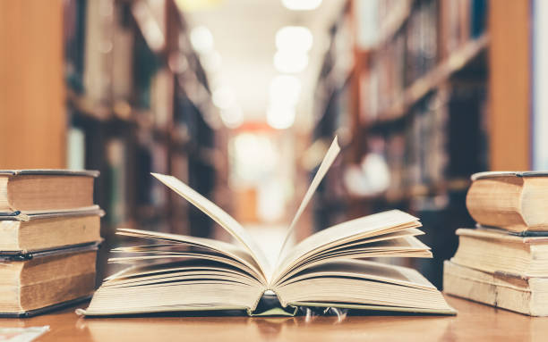 Education concept with book in library Book in library with old open textbook, stack piles of literature text archive on reading desk, and aisle of bookshelves in school study class room background for academic education learning concept encyclopaedia stock pictures, royalty-free photos & images