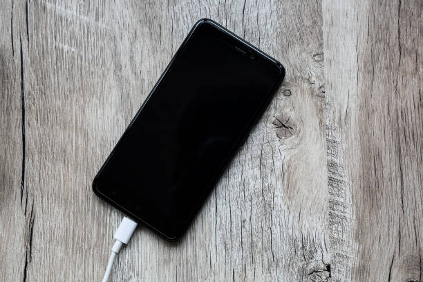 istock Black smartphone with white usb cable on wooden background. Modern gadget mobile phone with charger. Information technology. 944631342