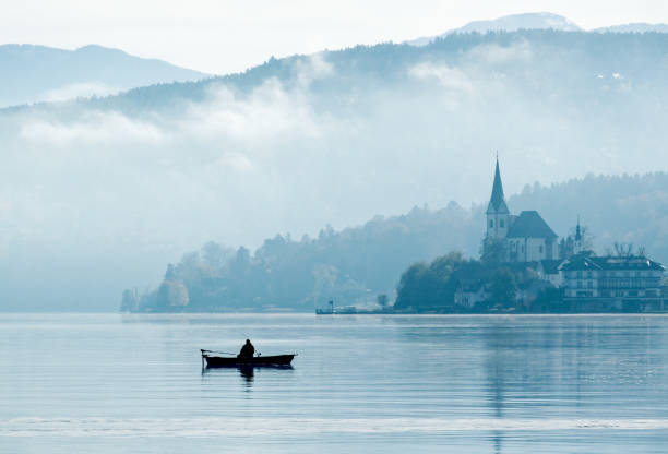 sylwetka samotnego rybaka siedzącego w łodzi wiosłowej z odbiciem jeziora w słońcu mglistego poranka - church steeple silhouette built structure zdjęcia i obrazy z banku zdjęć