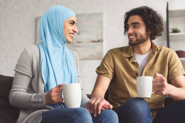sonriente pareja musulmana las manos y sosteniendo tazas de café en casa - parejas musulmanas fotografías e imágenes de stock