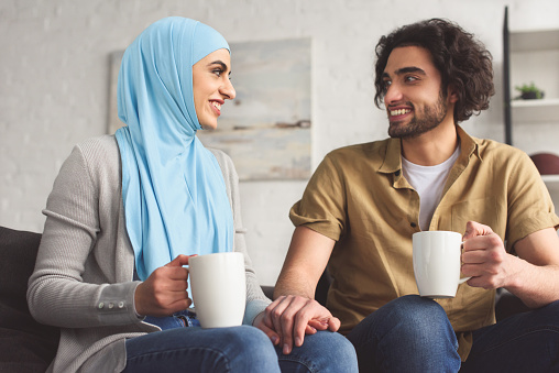 smiling muslim couple holding hands and holding cups of coffee at home