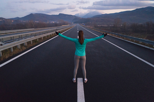 Rear view of a young female athlete standing on the highway with her arms outstretched.