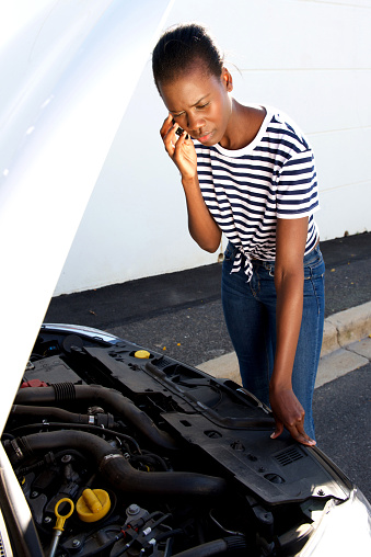 Portrait of upset young african woman standing by  broken down car parked on the side of a road and calling for assistance