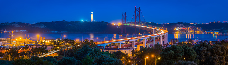 The iconic span of the 25 de Abril Bridge illuminated by the lights of the suspension cables and the zooming traffic crossing from Almada to Lisbon, Portugal’s vibrant capital city.