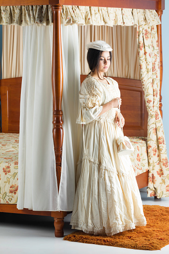A lady in her late 20s posing in a cream Victorian dress next to her four-poster bed.