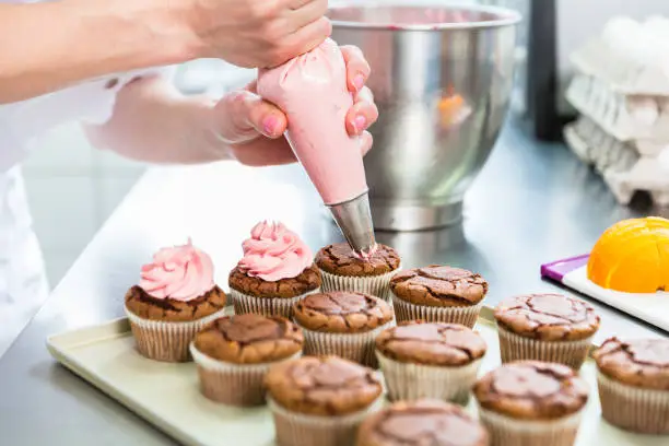 Women in pastry bakery as confectioner glazing muffins with icing bag, close-up