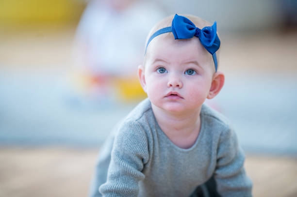 A Caucasian baby girl is indoors in a daycare center.  She is crawling and looking around curiously.