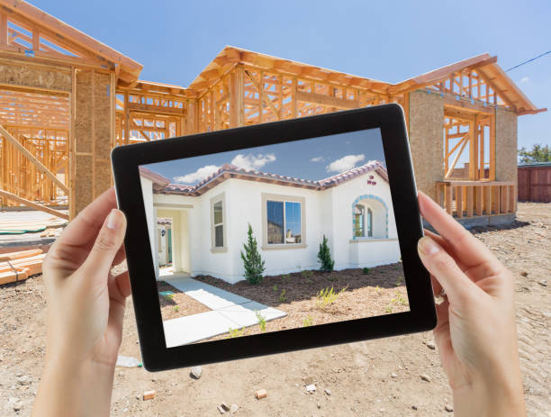 istock Female Hands Holding Computer Tablet with Finished House on Screen, Construction Framing Behind. 944751326