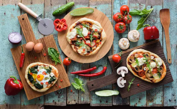 Flatlay of pizza party. Homemade pizzas and raw vegetables in abundance on oak chopping boards over shabby blue wooden background overhead view