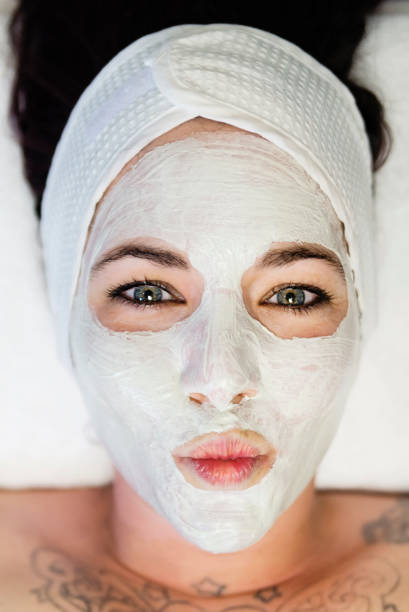 Young woman making a fund face with a beauty mask on. She is lying on her back and has a headband. She has some tattoos on her upper chest. Vertical indoors close-up high angle view shot with copy space.