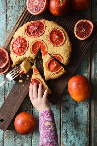 Baby taking piece of cake. Healthy homemade blood orange cake with cornmeal on dark oak chopping board over shabby rustic background overhead view