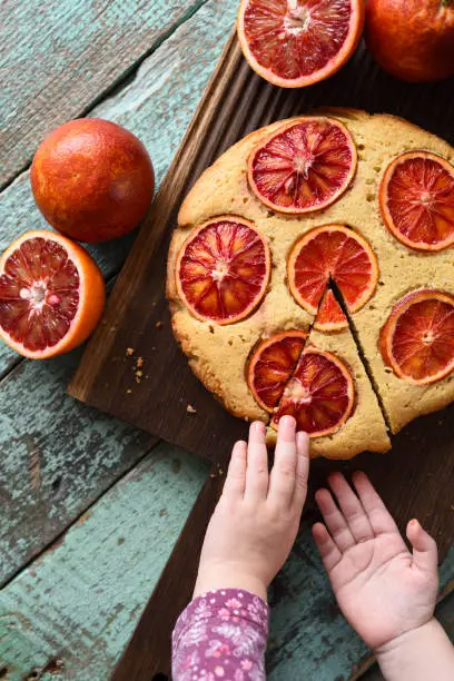 Dessert for children. Little girl hands reaching for piece of homemade cake with blood oranges on oak board top view