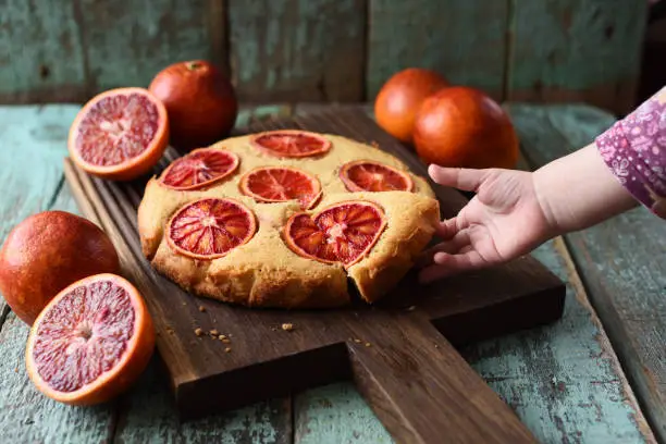 Sweets for children. Toddler hand reaching for homemade cake with blood orange slices on dark oak board on shabby background closeup