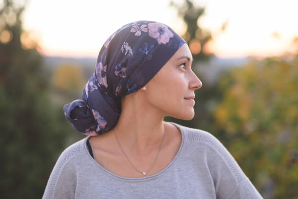 woman battling cancer stands outside and contemplates her life A beautiful young woman wearing a head wrap looks off-screen right and smiles radiantly. She is standing outdoors and there are mountains and trees in the background. survival stock pictures, royalty-free photos & images