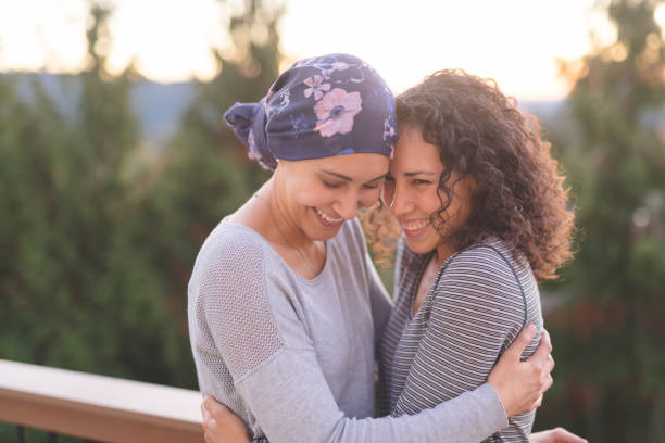 Beautiful woman battling cancer hugs her sister tightly A beautiful young woman fighting cancer and wearing a head wrap embraces her sister. They are tightly holding each other and she is looking down and smiling. Her sister is also smiling. They are standing outdoors and there are mountains and trees in the background. food chain stock pictures, royalty-free photos & images