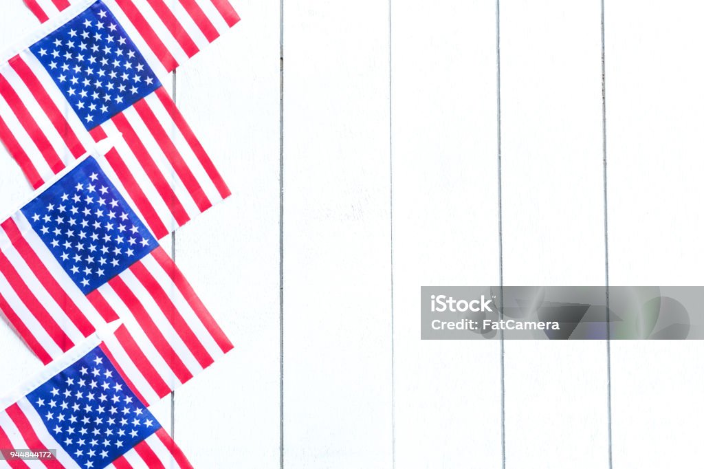 Stars and Stripes Forever A collection of American flags are displayed neatly in a row outside a home with white siding. They are in a vertical row, and the flags are at an angle hanging down from the upper left by the stars. American Culture Stock Photo