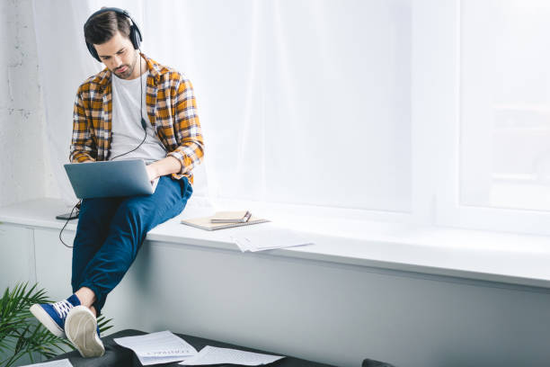 istock Man in headphones working on laptop and sitting on windowsill 944847612