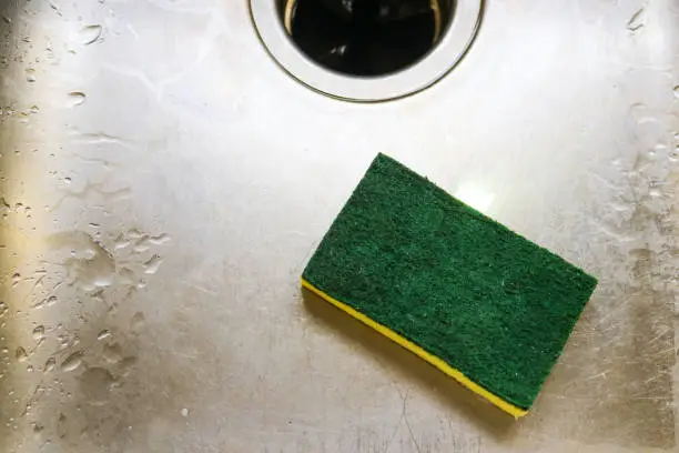 Green and yellow scrubing sponge lying in scratch and wet sink with garage disposal