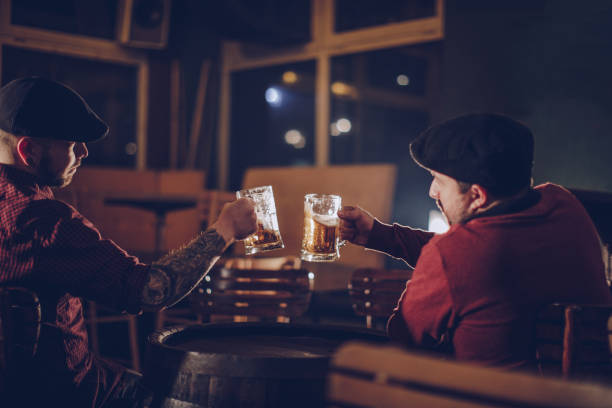 Two friends in pub drinking beer Two men in pub, men sitting and talking together in a pub. body adornment rear view young men men stock pictures, royalty-free photos & images