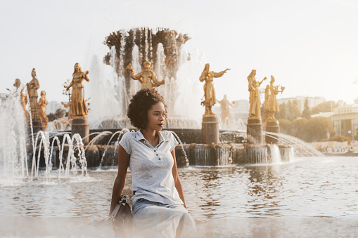 Pensive young glamorous African-American girl is sitting on the marble border of the fountain with a golden statues behind and thoughtfully looking aside, warm early summer evening